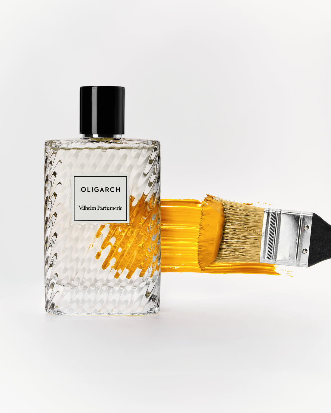 A clear textured glass perfume bottle labeled Oligarch - Villeso Parfumie with a black cap, shown beside a paintbrush with yellow-gold paint on a clean white background.